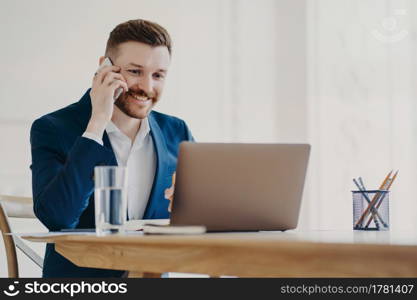 Young attractive businessman in formal suit smiling when talking on phone and looking at laptop screen approving what he sees, ceo sitting in minimalistic office environment with stylish work place. Young stylish executive talking on phone in front of laptop behind office desk