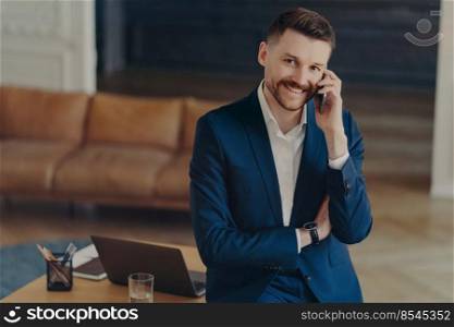 Young attractive businessman or male entrepreneur in formal dark blue suit talking on mobile phone with partner, looking happy and successful, leaning on desk while working from modern home or office. Smiling male entrepreneur talking on mobile phone while leaning on office desk