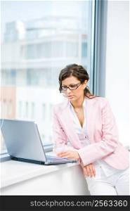 Young attractive businesswoman browsing internet on laptop computer in fron front of office window.