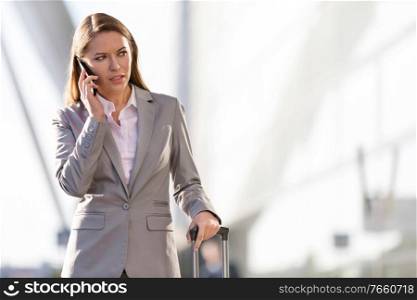 Young attractive businesswoman talking on smartphone while walking with her suitcase in airport