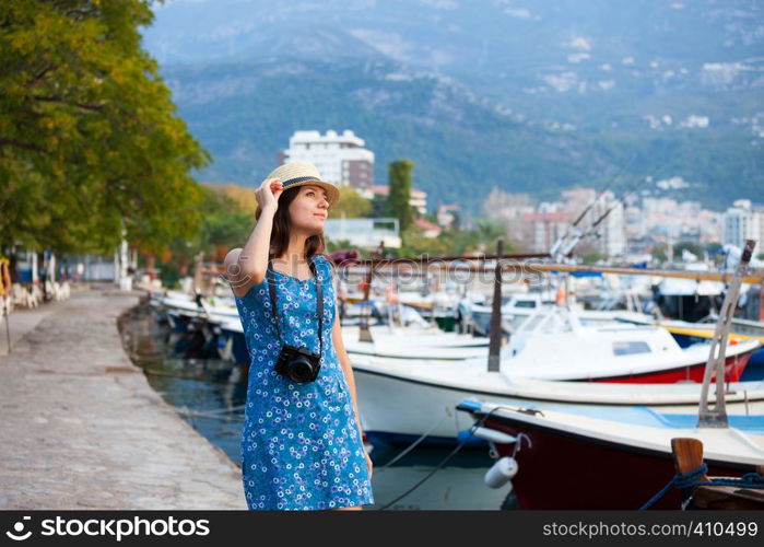 Young attractive cheerful travel girl walking with camera in old Mediterranean city seafront. Summer resort travel concept. Young attractive cheerful travel girl walking with camera