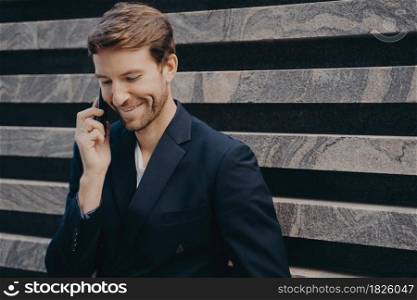 Young attractive confident businessman with neat beard in formal wear speaking on mobile leans against outer wall of city building. Smiling man having conversation on phone while having work break. Young attractive confident businessman with neat beard in formal wear speaking on mobile outdoors