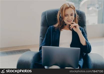 Young attractive european woman wearing headset sitting on armchair at home office, taking part in educational webinar, smiling female using laptop during video call with clients, working remotely. Young attractive european woman wearing headset sitting with laptop on armchair at home office