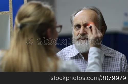 Young attractive female doctor examining senior man&acute;s eyesight with flashlight in medical office. Elderly man having reflex symptomatology checked by professional female neurologist in clinic.