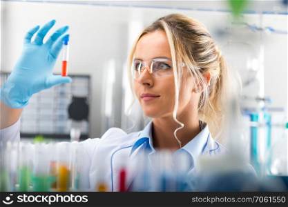 Young attractive female scientist in protective eyeglasses and gloves examining test tube with red liquid sample substance probe in the scientific chemical research laboratory