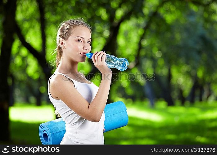 Young attractive girl drinking water and keeps the gym mat