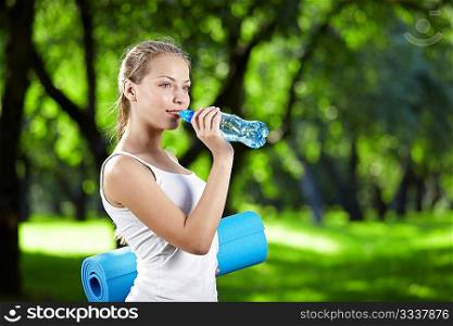 Young attractive girl drinking water and keeps the gym mat