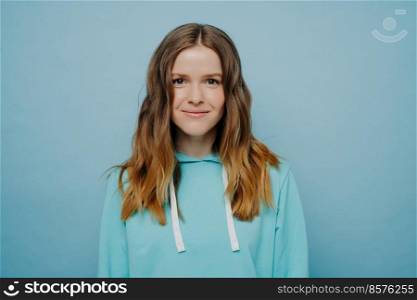 Young attractive girl with wavy ombre hairstyle smiling while looking at camera in comfortable casual hooded sweatshirt standing against light blue background. Medium shot. Portrait of teenage girl smiling at camera posing isolated over blue background