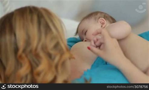 Young attractive mother breastfeeding her naked baby girl at home. Top angle view. Rear view. Caring mom stroking infant tiny arm and kissing her child with love and tenderness while breastfeeding indoors.