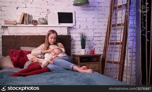 Young attractive mother caressing her beautiful daughter with tenderness while she is falling asleep on her knees in the bedroom. Cute little girl holding teddy bear and falling asleep on mother&acute;s knees.