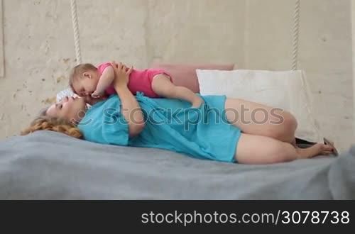 Young attractive mother lying on her back in bed, embracing her baby girl, gently kissing and playing while relaxing in bedroom at home. Sensual mother expressing her love and tenderness to little child.