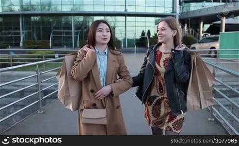 Young attractive multiracial girl friends with shopping bags holding hands walking on pedestrian bridge and chatting after shopping. Excited shopaholic females with packages discussing their purchases while walking on bridge in the city. Slow motion.