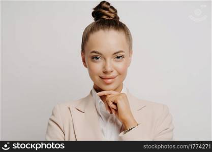 Young attractive self confident business woman in beige suit keeping hand under chin while posing on gray background being confident of herself, looking forward while thinking about profit or business. Assertive executive woman looking with confident look
