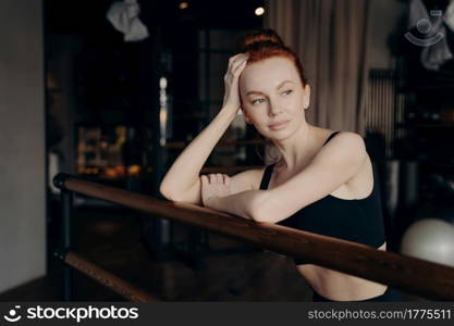 Young attractive slender woman ballerina with red hair leaning on ballet barre in meditative facial expression during stretching workout in fitness studio, resting and thinking about something. Young attractive woman with red hair stands leaning on ballet barre and resting after workout