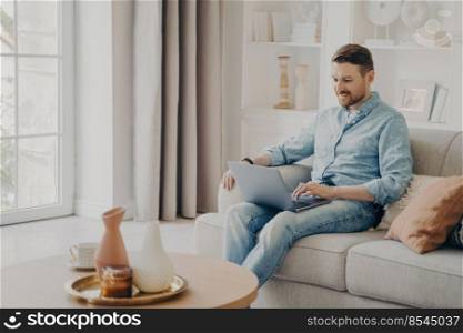 Young attractive smiling male freelancer sitting on comfortable beige sofa at home, using laptop and checking his emals while working remotely during weekend, wearing casual clothes. Young attractive smiling freelancer working on comfortable sofa at home
