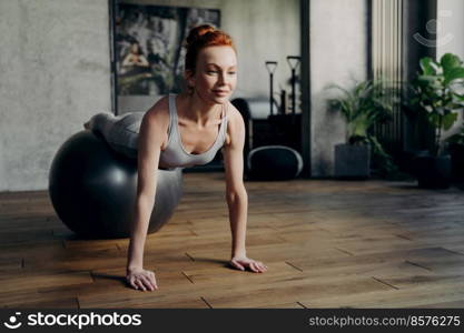 Young attractive smiling red haired woman training in fitness studio, lying on exercise ball and doing push ups with straight arms based on floor, sportive motivated female working out with fitball. Fit woman balancing on exercise ball in fitness studio during pilates workout