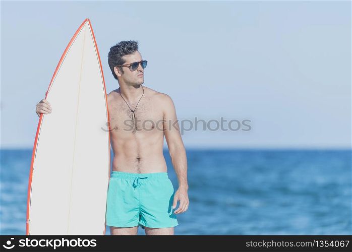 Young attractive surfer holding his surfboard at the beach wearing a green swimsuit