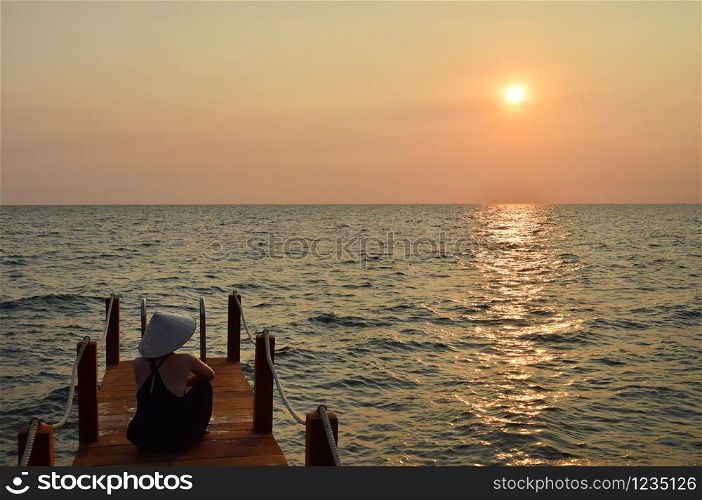 Young attractive woman at the seashore in Vietnam wearing a typical hat staring at the sunset.. Lonely girl watching the sunset in Vietnam.
