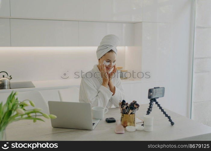 Young attractive woman blogger applying cosmetic patches under eyes recording video on smartphone for beauty vlog, creating content for social media while sitting in modern kitchen. Skin care concept. Female beauty blogger in bathrobe and towel on head recording video on smartphone for her vlog