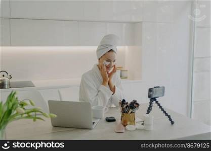 Young attractive woman blogger applying cosmetic patches under eyes recording video on smartphone for beauty vlog, creating content for social media while sitting in modern kitchen. Skin care concept. Female beauty blogger in bathrobe and towel on head recording video on smartphone for her vlog