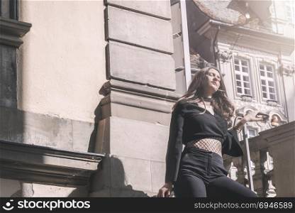 Young attractive woman dressed in black modern clothes, leaning against a balustrade, surrounded by aged German architecture, enjoying the sun.