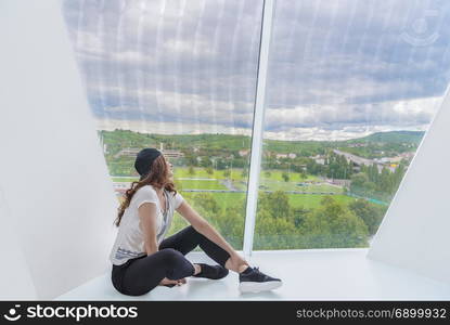 Young attractive woman, dressed in casual clothing, sits on a white floor from a modern building and looks through a very tall window.