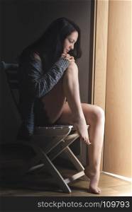 Young attractive woman dressed sexy and cozy, sitting on a chair with her head leaned against her naked feet, meditating, in front of an open door.