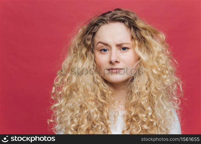Young attractive woman has discontent expression, raises eyebrow in bewilderment, being dissatisfied with something, has bushy curly hair, isolated over pink background. Facial expressions concept