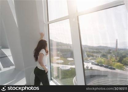 Young, attractive woman, in a modern building, dressed casual, looking forward, on a large window, with the sun shining over her.