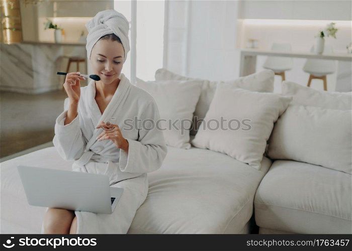 Young attractive woman in bathrobe and towel on head applying powder on face while watching make up video tutorial on laptop, sitting on sofa in modern living room at home and getting ready for work. Young pretty woman in bathrobe applying powder on face while doing daily makeup at home