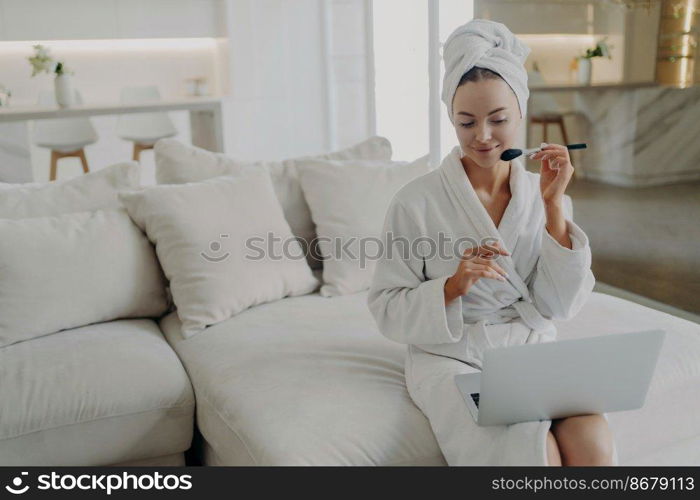Young attractive woman in bathrobe and towel on head applying powder on face while watching make up video tutorial on laptop, sitting on sofa in modern living room at home and getting ready for work. Young pretty woman in bathrobe applying powder on face while doing daily makeup at home
