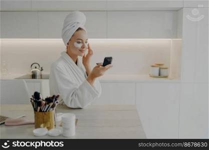 Young attractive woman in bathrobe and towel turban on head applying cosmetic patches under eyes from dark circles, looking in compact mirror and smiling while standing in modern kitchen at home. Happy woman in white bathrobe with towel on head applying cosmetic patches under eyes