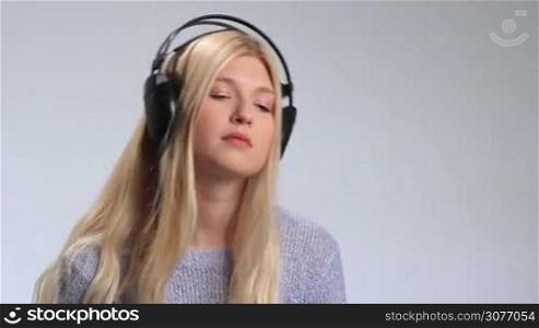 Young attractive woman in big black headphones listening to melancholic music closeup on white. Teenager girl moving slowly to sad song, looking at camera with sad facial expression, closing her eyes and continues listening to music in headphones.