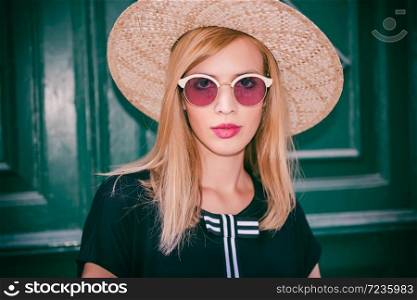 Young attractive woman in fashionable black dress,and stylish round sunglasses and yellow hat poses in front of wall at street.Retro and vintage filter and colors effect used.. Young attractive woman poses in front of wall