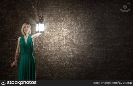 Young attractive woman in green dress with lantern walking in darkness. Light of education