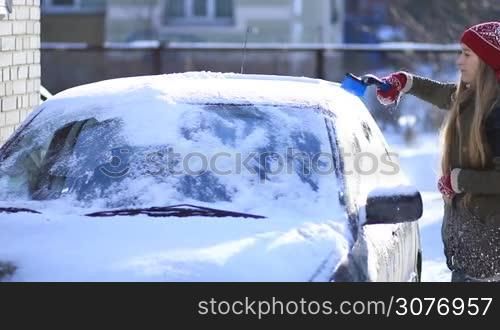 Young attractive woman in red woolen gloves and hat removing snow from car&acute;s roof with the help of special snow brush. Female cleaning car from snow on winter morning.