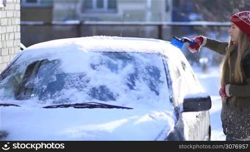 Young attractive woman in red woolen gloves and hat removing snow from car&acute;s roof with the help of special snow brush. Female cleaning car from snow on winter morning.