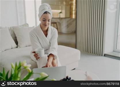 Young attractive woman in white bathrobe and hair wrapped in towel relaxing on sofa in living room after taking shower or bath at home, happy female drinking tea or coffee, using laptop and smiling. Young woman in white bathrobe working remotely on laptop while sitting on sofa in modern living room