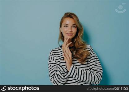 Young attractive woman poses with hands folded gently touches her face and looks tenderly at camera with slightly open mouth, standing isolated over light blue background dressed in striped longsleeve. Young attractive woman with hands folded on chest gently touching her face and smiling at camera