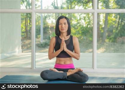 Young attractive woman practicing yoga, sitting in Padmasana, exercise, Lotus pose, namaste, working out, wearing sportswear,  pants black, indoor full length, near floor window.