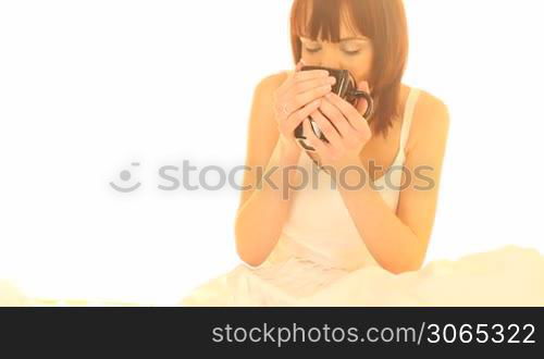 Young attractive woman seated on her bed enjoying a large mug of morning coffee