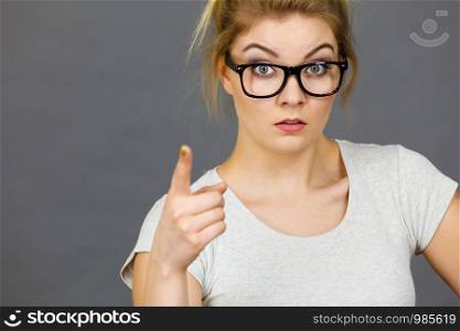 Young attractive woman wearing eyeglasses pointing into camera, teenage judging someone. Studio shot on grey background.. Woman wearing eyeglasses pointing at camera