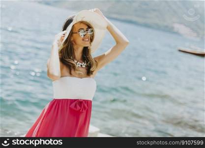 Young attractive woman with a hat and sunglasses poses by the sea at sunny day