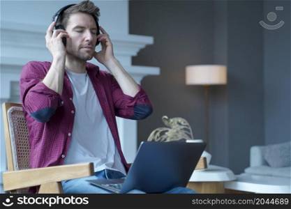 Young austrian guy sitting on wooden wicker chair with open laptop on his lap listening music in wireless headphones totally immersed in favorite, sounds closing eyes from pleasure of song melodies. Young german guy sitting on wicker chair with laptop on his lap listening music in headphones