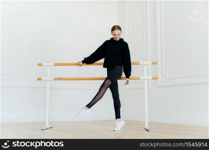 Young ballet dancer has warm up, stands near handrails, dressed in black sportclothes, stands on tip toe, wears white sneakers, focused down on floor, has cheerful facial expression, raises leg