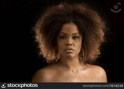 Young bare shouldered African American female model with curly hair and elegant makeup with golden hoop earrings and necklace looking at camera against black background. Attractive curly haired black woman with makeup