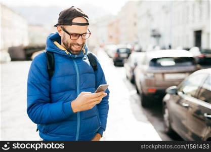 Young bearded hipster guy in blue anorak and cap holding smartphone answering call having happy expression isolated over big city background. Stylish man using cell phone while standing on street