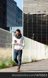 Young bearded male with headphones and holding smartphone while walking against skyscrapers in sunny day