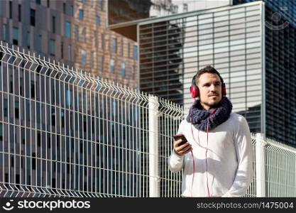 Young bearded male with headphones and holding smartphone while walking against skyscrapers in sunny day