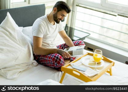 Young bearded  man having breakfast in bed and reading newspaper in the room